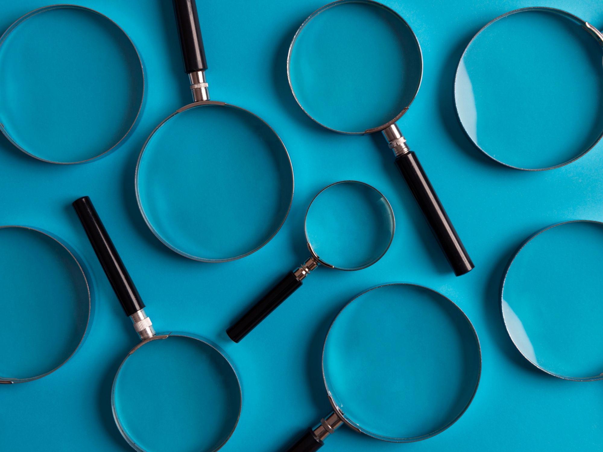 Several magnifying glasses of different sizes with black handles and silver rims arranged on a bright blue background.