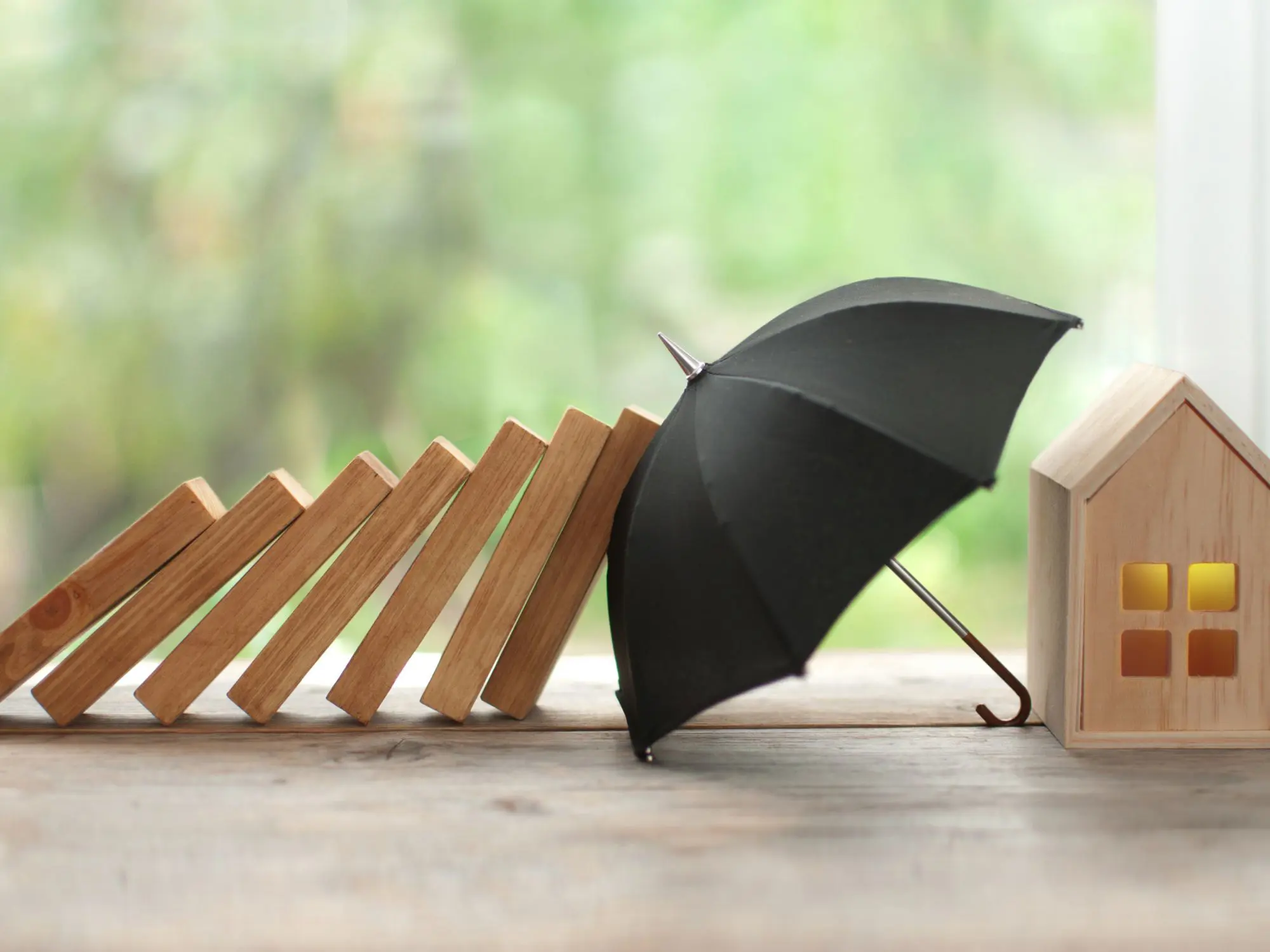 A row of falling wooden dominoes heads toward a small black umbrella shielding a wooden house with yellow windows, set against a blurred green background.