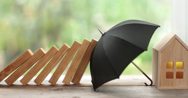 A row of falling wooden dominoes heads toward a small black umbrella shielding a wooden house with yellow windows, set against a blurred green background.