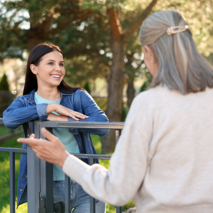 Two people talking over a low metal fence in a green outdoor setting, with trees and sunlight in the background. One person is leaning on the fence while listening, and the other is facing them with a hand gesture, suggesting a conversation between neighbours. Provide your feedback on BizChat