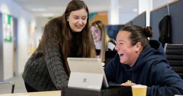 Three people collaborating in a modern office or study space; two are focused on a laptop, with one standing and the other seated and smiling, while a third person works independently in the background.