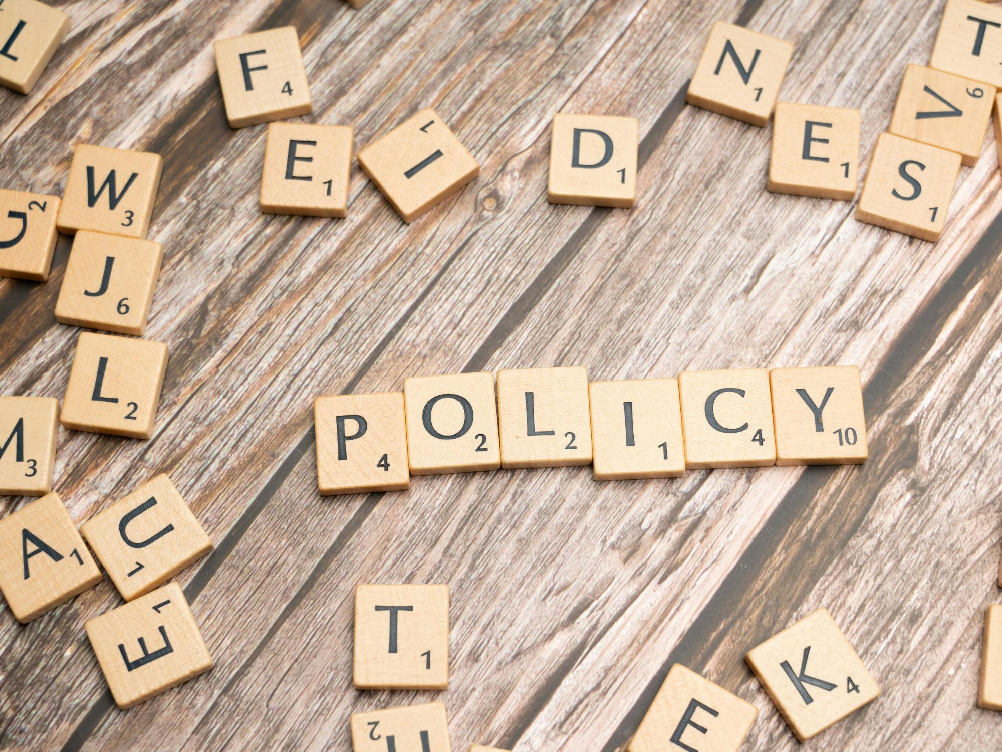 Scrabble tiles on a wooden surface spell out the word "POLICY" in the centre, with other random tiles scattered around.
