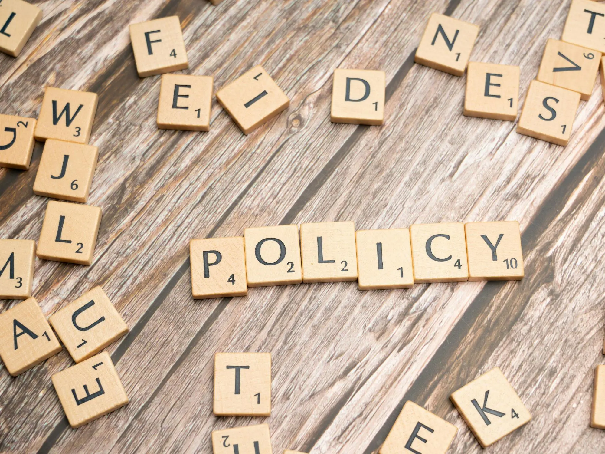 Scrabble tiles on a wooden surface spell out the word "POLICY" in the centre, with other random tiles scattered around.