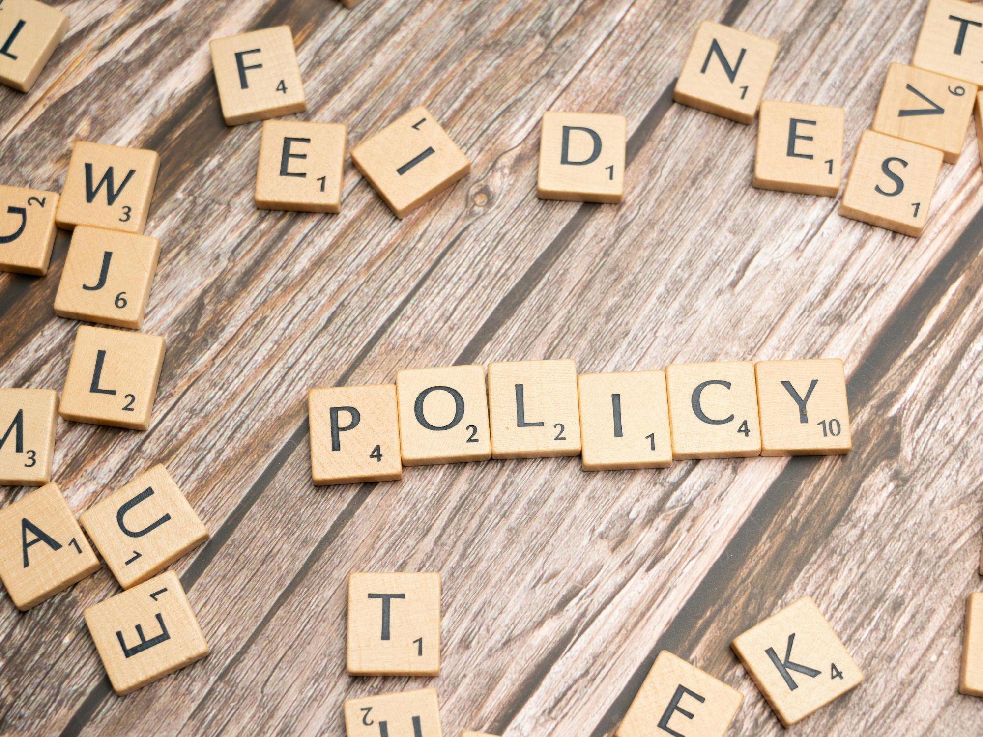 Scrabble tiles on a wooden surface spell out the word "POLICY" in the centre, with other random tiles scattered around.