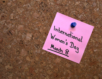 “Pink sticky note pinned to a corkboard with a blue pushpin, handwritten with the words ‘International Women’s Day, March 8’.”