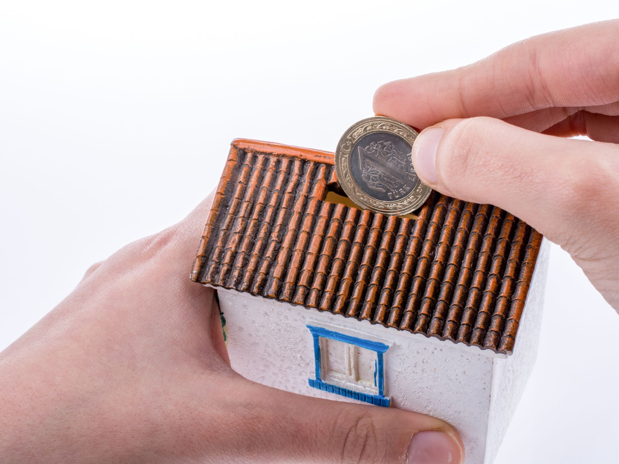 A person inserts a coin into the slot of a small house-shaped piggy bank with a tiled roof, white walls, and a blue-framed window, symbolising saving for home ownership.