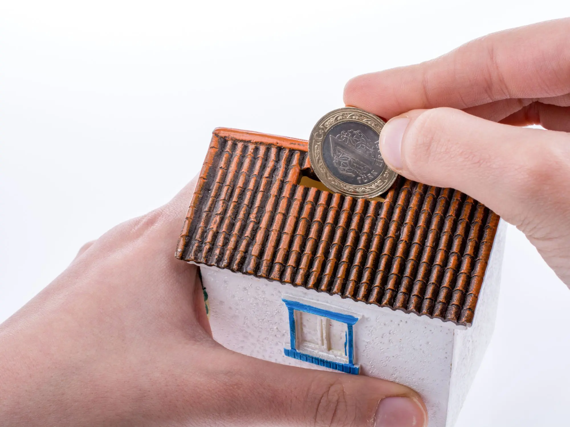 A person inserts a coin into the slot of a small house-shaped piggy bank with a tiled roof, white walls, and a blue-framed window, symbolising saving for home ownership.