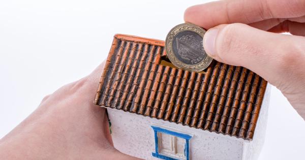A person inserts a coin into the slot of a small house-shaped piggy bank with a tiled roof, white walls, and a blue-framed window, symbolising saving for home ownership.