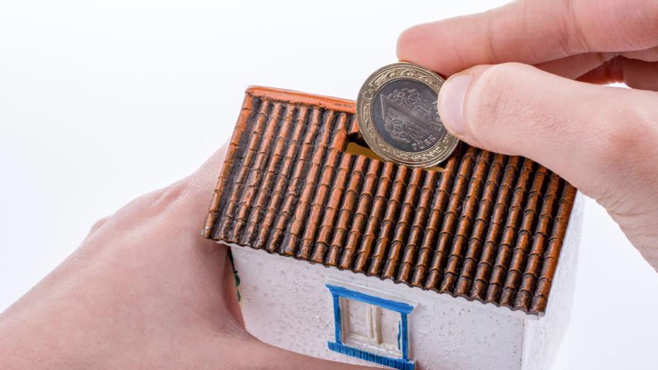 A person inserts a coin into the slot of a small house-shaped piggy bank with a tiled roof, white walls, and a blue-framed window, symbolising saving for home ownership. A person inserts a coin into the slot of a small house-shaped piggy bank with a tiled roof, white walls, and a blue-framed window, symbolising saving for home ownership.
