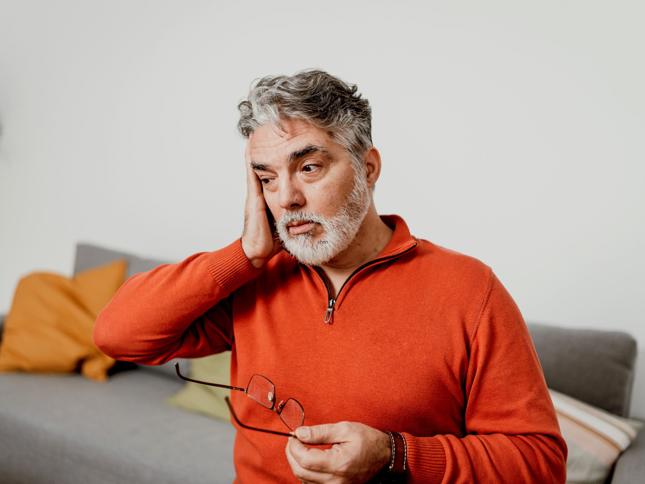 An older person with gray hair and a beard, holding glasses and touching their head, appearing thoughtful or concerned. An older person with gray hair and a beard, holding glasses and touching their head, appearing thoughtful or concerned.