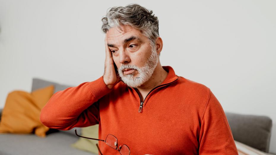 An older person with gray hair and a beard, holding glasses and touching their head, appearing thoughtful or concerned. An older person with gray hair and a beard, holding glasses and touching their head, appearing thoughtful or concerned.
