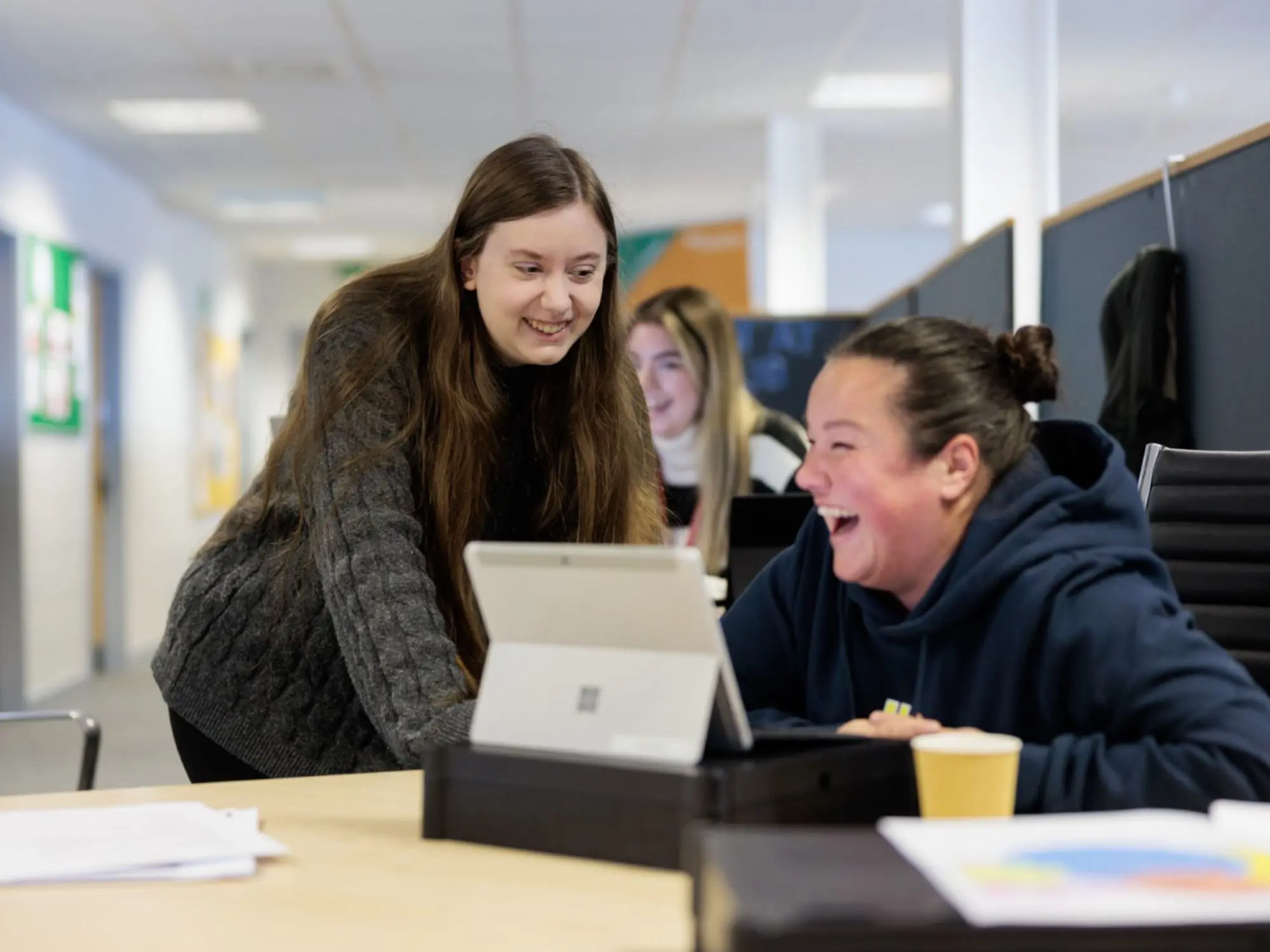Two people in a collaborative office setting, one standing and leaning over a table to look at a laptop, while the other is seated and laughing. Papers and a cup are on the table, with another person working at a computer in the background.