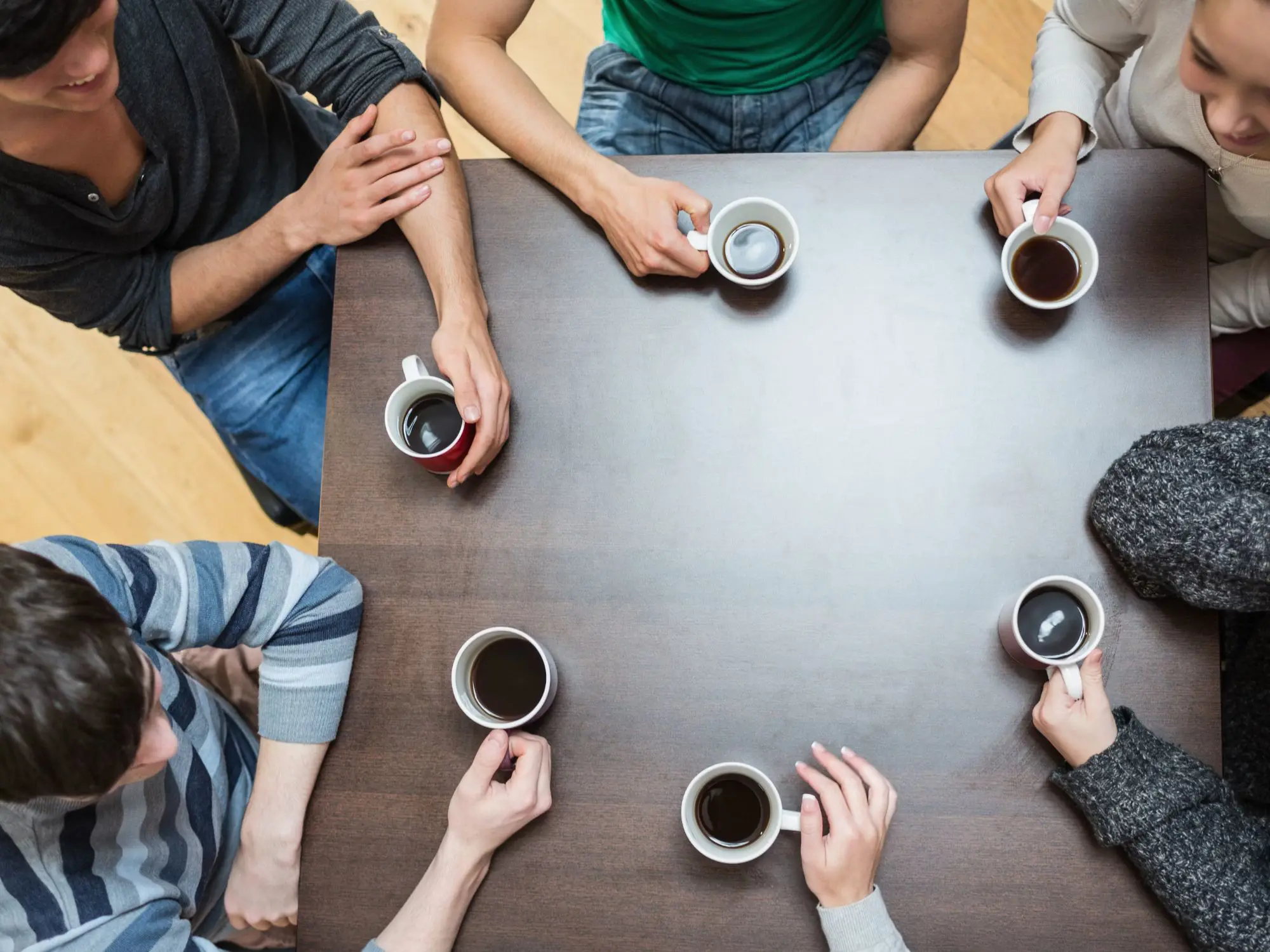 Six people seated around a dark wooden table, each holding a white mug filled with a dark beverage, likely coffee or tea. The mugs are arranged in a loose circle, and the individuals’ arms and hands are visible resting on or near the table. The setting appears casual, with a light wooden floor in the background.