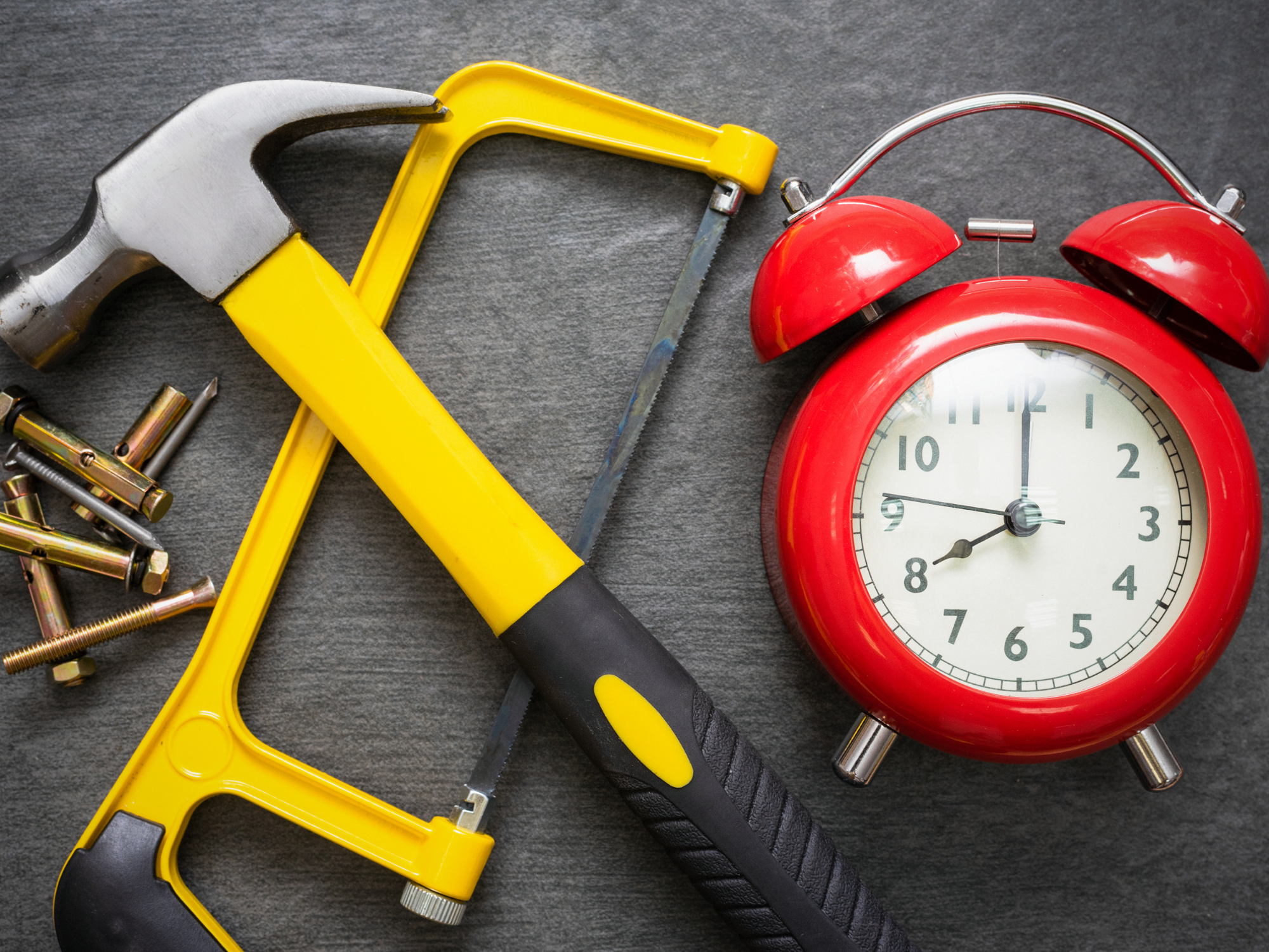 Assorted tools including a hammer, hacksaw, screws, bolts, and a red alarm clock arranged on a dark surface.