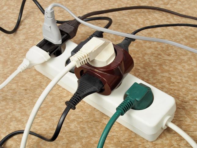 White power strip with six occupied outlets, featuring various colored plugs and adapters, placed on a beige carpeted floor. White power strip with six occupied outlets, featuring various colored plugs and adapters, placed on a beige carpeted floor.