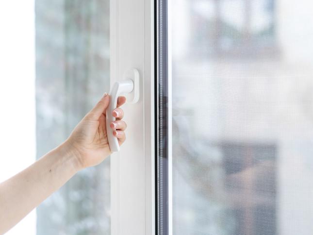 Hand turning a white handle on a partially open window with a mesh screen, and blurred buildings visible in the background. Hand turning a white handle on a partially open window with a mesh screen, and blurred buildings visible in the background.