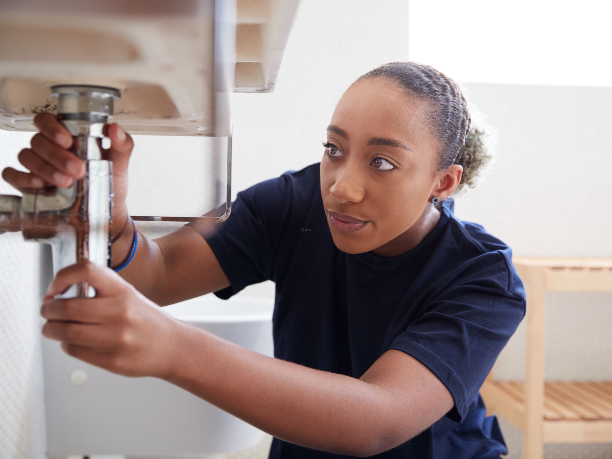Person working under a bathroom sink, adjusting pipes with both hands, with a toilet and wooden shelf visible in the background.