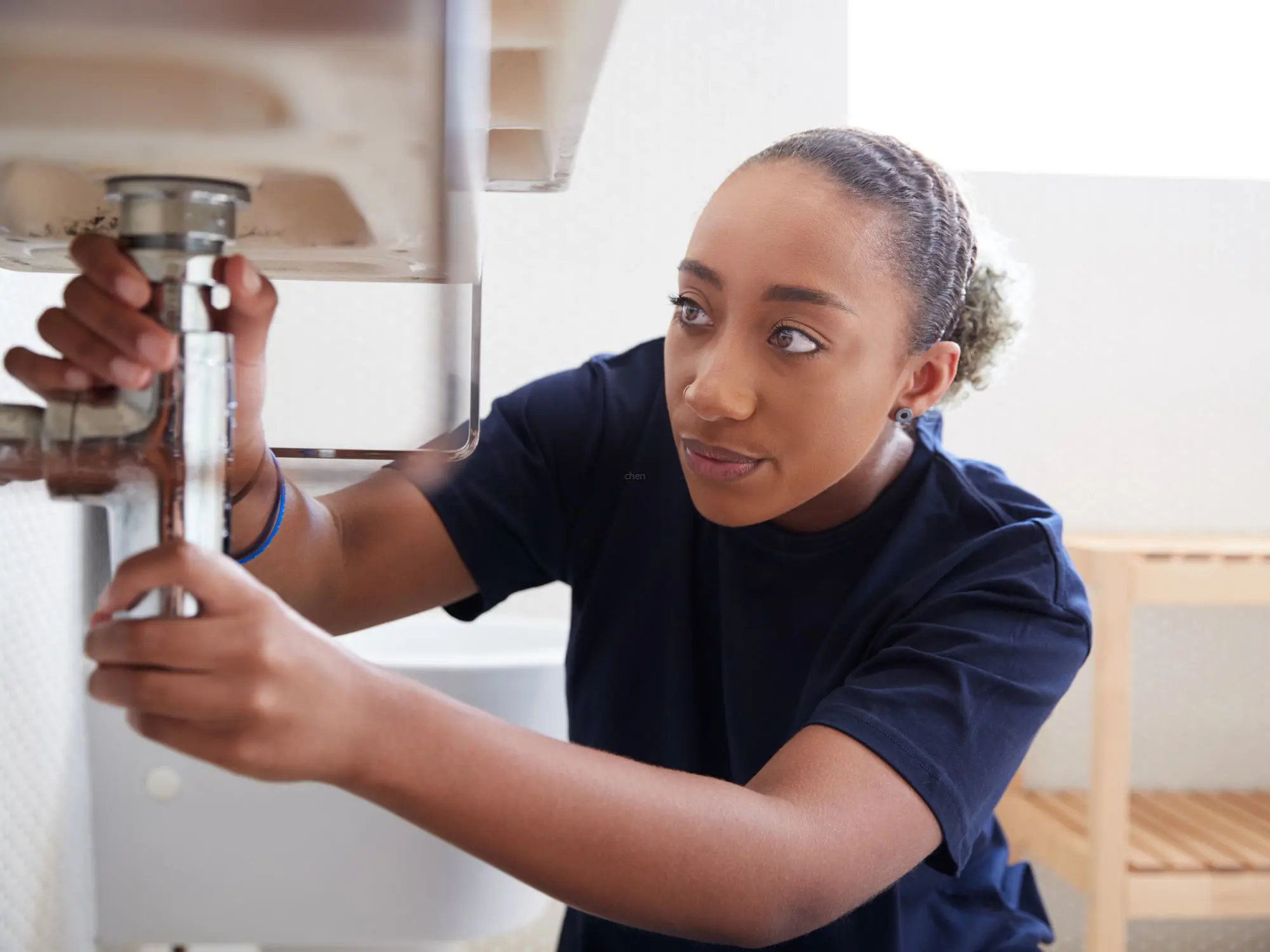 Person working under a bathroom sink, adjusting pipes with both hands, with a toilet and wooden shelf visible in the background.