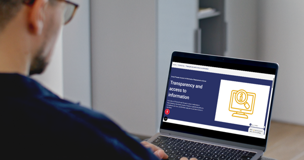 Person using a laptop to view a webpage about transparency and access to information. The screen displays a blue information page with an accessibility icon, while the person sits in a home or office setting.