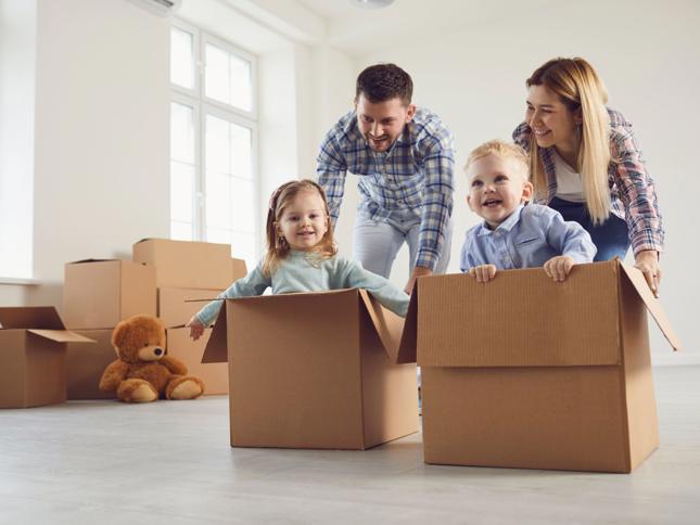Two children sitting inside cardboard boxes while two adults playfully push them across the floor in a bright room with moving boxes and a teddy bear in the background. Two children sitting inside cardboard boxes while two adults playfully push them across the floor in a bright room with moving boxes and a teddy bear in the background.