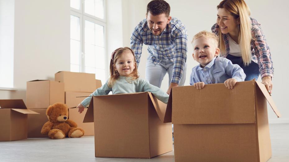 Two children sitting inside cardboard boxes while two adults playfully push them across the floor in a bright room with moving boxes and a teddy bear in the background. Two children sitting inside cardboard boxes while two adults playfully push them across the floor in a bright room with moving boxes and a teddy bear in the background.