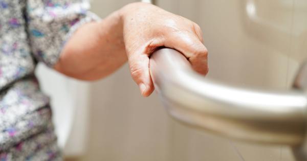 Close-up of an elderly person's hand gripping a stainless steel grab bar in a bathroom, with a toilet and tiled wall in the background.