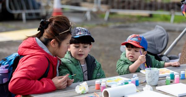 An adult and two children sit at an outdoor wooden table covered with art supplies, including paint bottles, brushes, paper, and a cup of water. The group appears to be engaged in a painting activity, with colorful paints arranged neatly on the table. In the background, there are traffic cones, fencing, and a stroller, suggesting the setting is an open community space or garden area.