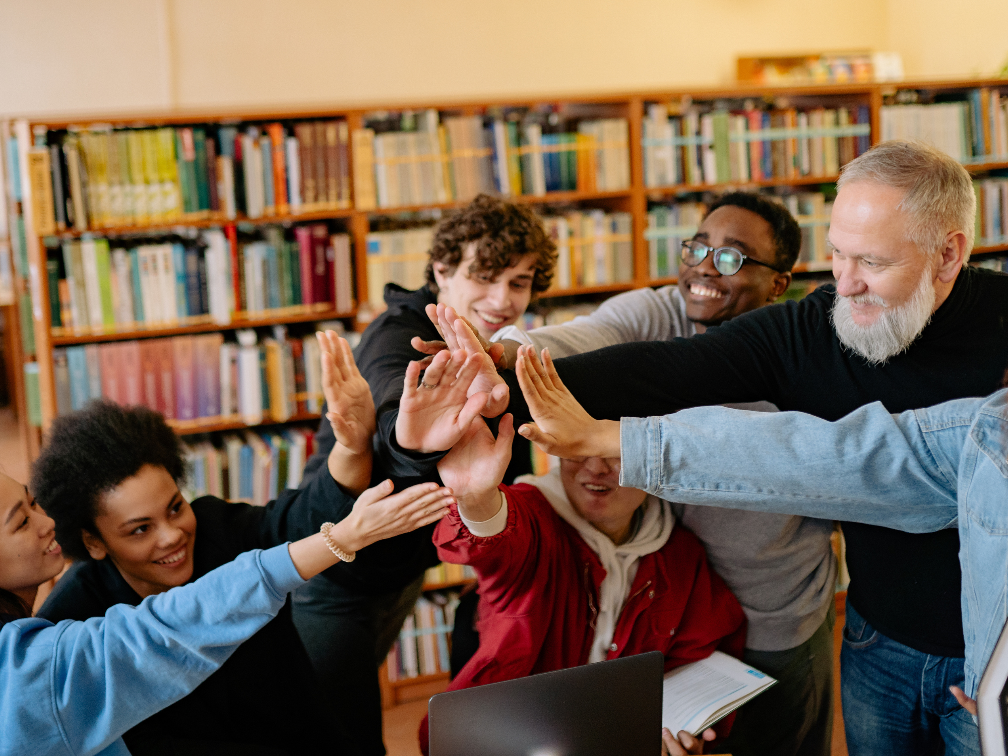 “Group of people seated around a table, reaching in to high‑five each other, with bookshelves in the background.”