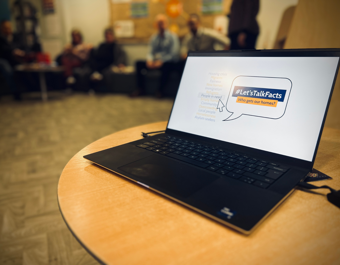 “A laptop on a round wooden table displays a slide with the hashtag ‘#Let’sTalkFacts – Who gets our homes?’ In the background, several people sit in a community‑room setting, out of focus.”