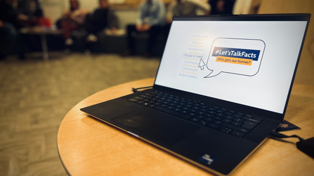 “A laptop on a round wooden table displays a slide with the hashtag ‘#Let’sTalkFacts – Who gets our homes?’ In the background, several people sit in a community‑room setting, out of focus.”