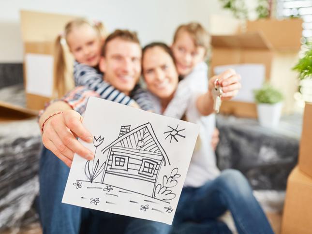 Person holding a drawing of a house in the foreground while another hand holds a set of keys, with moving boxes and plants in the background. Person holding a drawing of a house in the foreground while another hand holds a set of keys, with moving boxes and plants in the background.