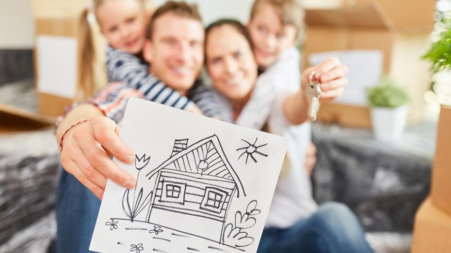 Person holding a drawing of a house in the foreground while another hand holds a set of keys, with moving boxes and plants in the background. Person holding a drawing of a house in the foreground while another hand holds a set of keys, with moving boxes and plants in the background.