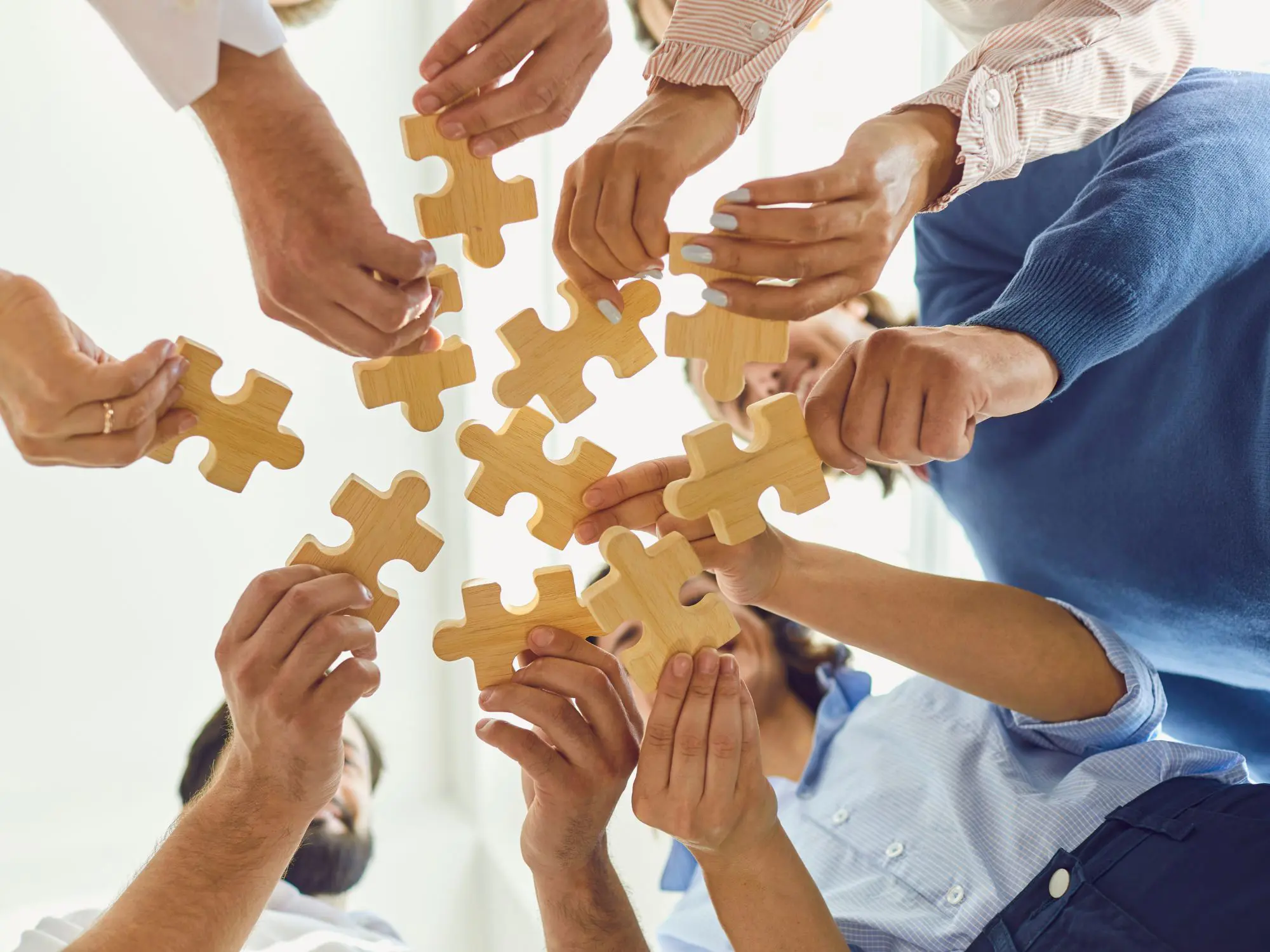 A group of people standing in a circle, each holding a large wooden puzzle piece, reaching toward the center to fit them together — symbolizing teamwork and collaboration.