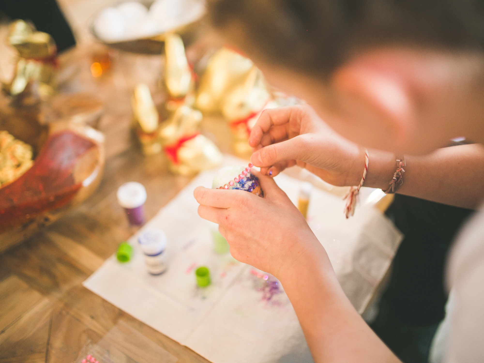 Close-up of a person decorating a craft item with colourful beads and glitter at a table covered with craft supplies, including paint pots and festive decorations.