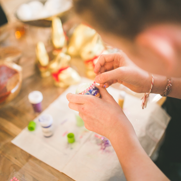 Close-up of a person decorating a craft item with colourful beads and glitter at a table covered with craft supplies, including paint pots and festive decorations.