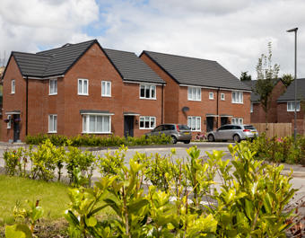 Row of modern red-brick townhouses with grey roofs, white-framed windows, front gardens, and parked vehicles in a suburban setting under a partly cloudy sky