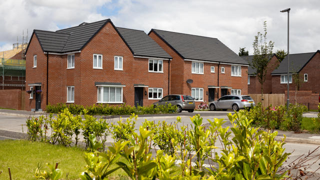 Row of modern red-brick townhouses with grey roofs, white-framed windows, front gardens, and parked vehicles in a suburban setting under a partly cloudy sky