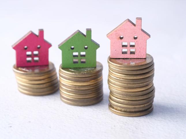 Three small wooden house models in red, green, and pink placed on stacks of coins of varying heights, arranged in a row to suggest different values or amounts. Three small wooden house models in red, green, and pink placed on stacks of coins of varying heights, arranged in a row to suggest different values or amounts.