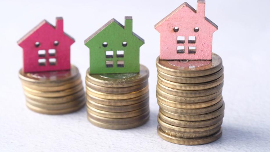 Three small wooden house models in red, green, and pink placed on stacks of coins of varying heights, arranged in a row to suggest different values or amounts. Three small wooden house models in red, green, and pink placed on stacks of coins of varying heights, arranged in a row to suggest different values or amounts.