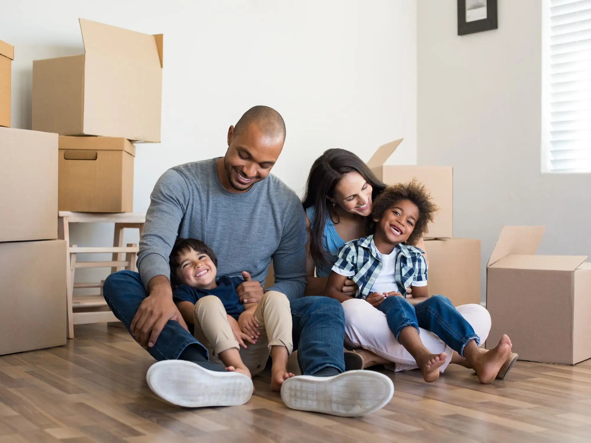A casually dressed family of four sits on the wooden floor of a light-colored room, surrounded by cardboard moving boxes. A window with blinds lets in natural light, suggesting they are moving into or out of a new home.