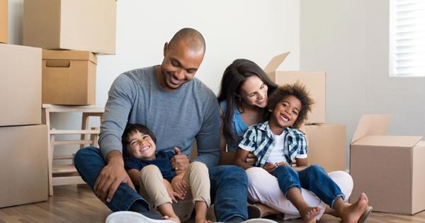 A casually dressed family of four sits on the wooden floor of a light-colored room, surrounded by cardboard moving boxes. A window with blinds lets in natural light, suggesting they are moving into or out of a new home.