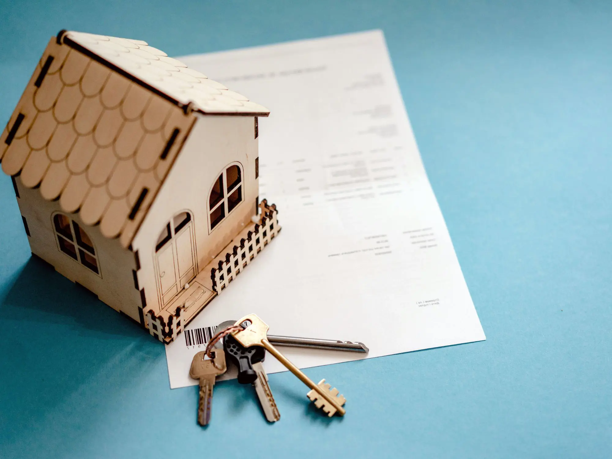 A small wooden house model with a picket fence sits atop a document, with several keys on a keyring placed in front. The background is a light blue surface, suggesting themes of property ownership or conveyancing.