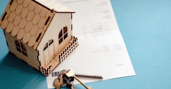 A small wooden house model with a picket fence sits atop a document, with several keys on a keyring placed in front. The background is a light blue surface, suggesting themes of property ownership or conveyancing.