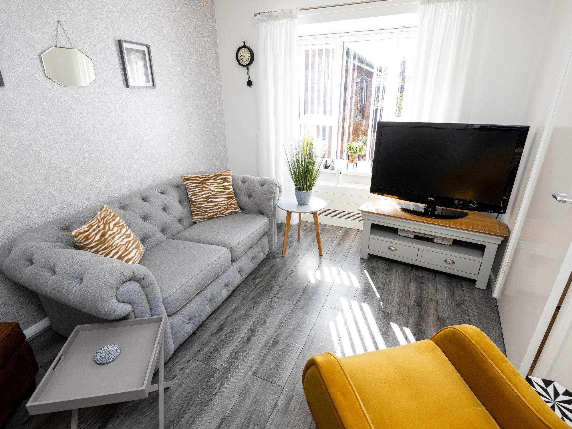 Modern living room with a grey tufted sofa, zebra-patterned pillows, a yellow armchair, and a wooden TV stand. A round table with a potted plant and decorative wall items complete the space.