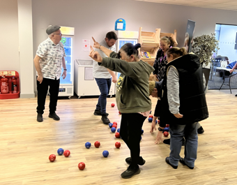 People taking part in a boccia activity indoors, with red and blue balls scattered across a wooden floor as participants reach down and move between them in a community space.