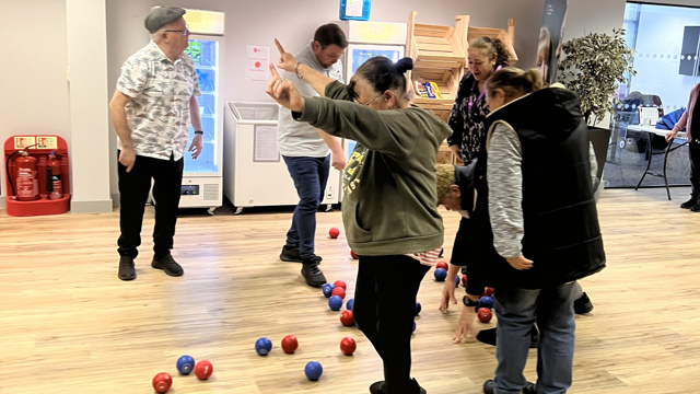 People taking part in a boccia activity indoors, with red and blue balls scattered across a wooden floor as participants reach down and move between them in a community space.