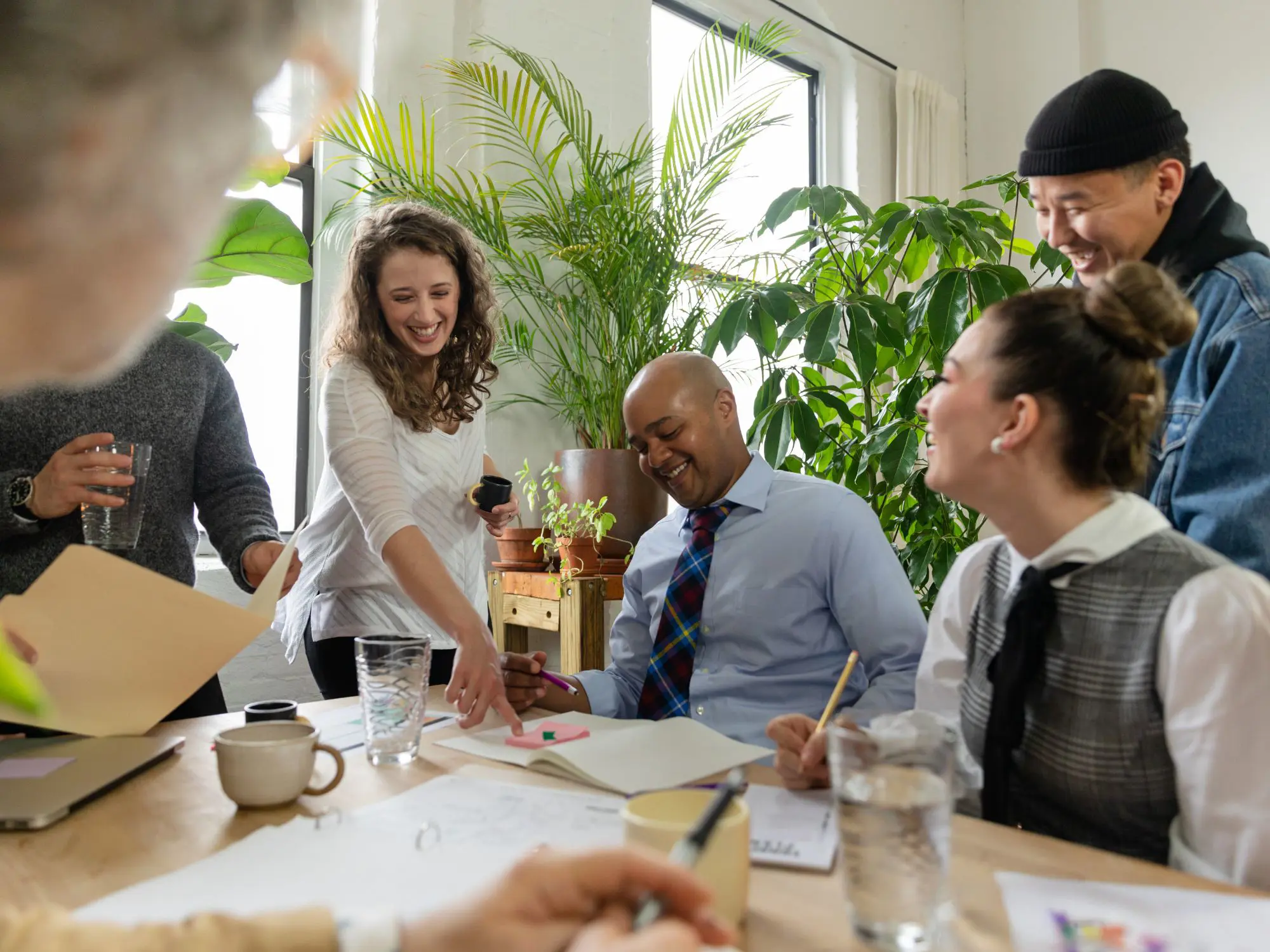 A group of people collaborating around a table in a bright office setting, with papers, notebooks, and glasses of water visible. Green plants decorate the space, adding a lively atmosphere.