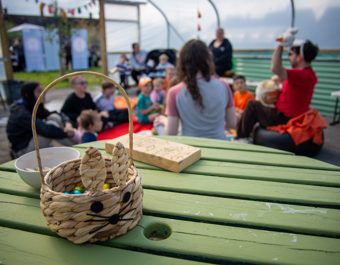 A small woven basket shaped like a bunny, filled with colourful sweets, sits on a green wooden table in the foreground. Behind it, a group of children and adults sit together on blankets and chairs inside an outdoor covered space at a community garden. Some participants appear to be taking part in an Easter‑themed activity, with one person holding up an item for the group. The setting is bright and open, with decorations and greenery visible in the background.