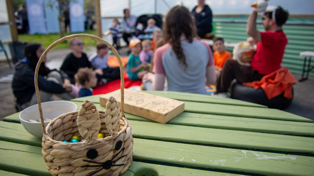 A small woven basket shaped like a bunny, filled with colourful sweets, sits on a green wooden table in the foreground. Behind it, a group of children and adults sit together on blankets and chairs inside an outdoor covered space at a community garden. Some participants appear to be taking part in an Easter‑themed activity, with one person holding up an item for the group. The setting is bright and open, with decorations and greenery visible in the background.