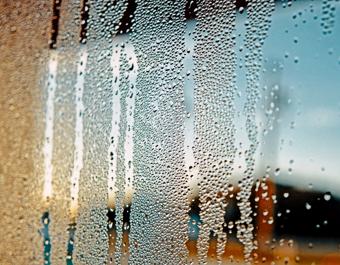 Close-up of a window with condensation, showing water droplets streaking down the glass. The blurred background reveals an outdoor scene with blue sky and faint structural or landscape elements.